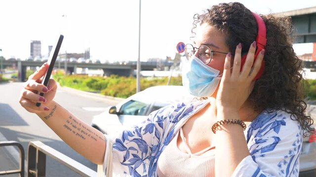 Young mixed race woman wearing surgical mask outdoor doing video call holding smartphone and with wireless bluetooth headphones