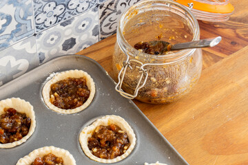 Mince pies in a baking tray with mincemeat in a clip glass jar and a spoon.  On a wood worktop with a tiled background