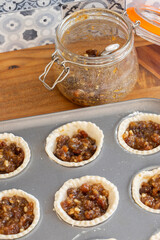 Mince pies in a baking tray with mincemeat in a clip glass jar and a spoon.  On a wood worktop with a tiled background
