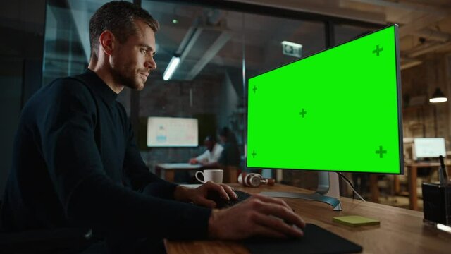 Young Handsome Specialist Working on Desktop Computer with Green Screen Mock Up Display in a Busy Creative Office with Colleagues. Male Manager with Trimmed Beard is Wearing a Casual Black Jumper.