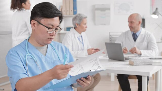 Medium close-up POV of young Asian male doctor sitting in medical office, leafing over docs in foreground of diverse colleagues talking and working at table. Man looking up and smiling on camera