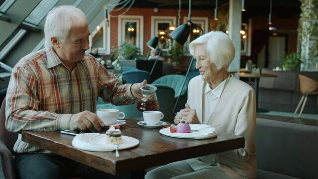 Senior man pouring tea in wife's cup taking care of woman talking and smiling in cafe, delicious desserts are visible on table. People and restaurants concept.