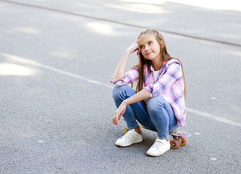 Smiling cute little girl child sitting with a skateboard. Preteen with penny board outdoors