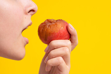 The girl bites a rotten apple with a worm on a yellow background. Expired products, junk food