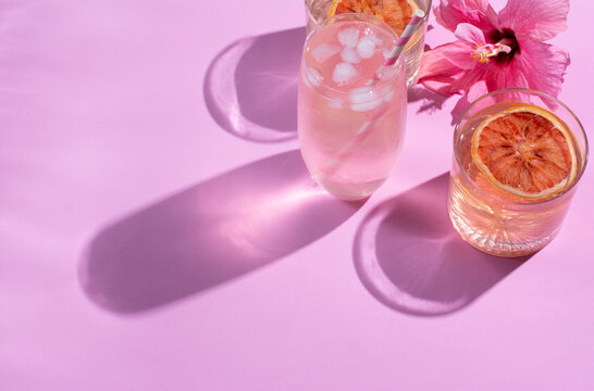Summer refreshing drink in glasses of water or cocktails and sliced fruits on a pink table background in sunlight. Long, sharp shadows. High angle. Lighting, leisure concept. A flower decorating