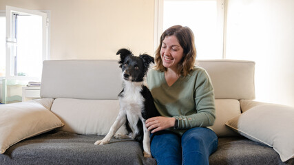 Smiling young woman sitting on the couch hugging a happy border collie puppy. Dog sitting next to young woman