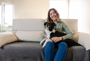 Smiling young woman sitting on the couch hugging a happy border collie puppy.