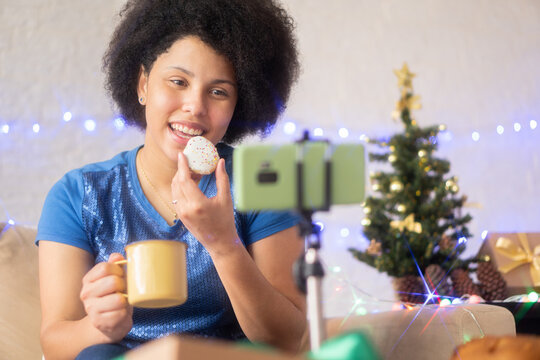 Person In Video Call With Distant Family And Friend At Christmas. Black Woman Eating Cookies And Drinking Eggnog Or Coffee In Celebration Of The Holiday Season.