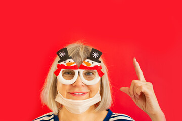 Fototapeta na wymiar Merry Christmas, Happy Holidays and New Year. Senior woman smiling thumbs up, wearing medical mask and Xmas glasses on isolated red background. Coronavirus quarantine lockdown. Stay at home.