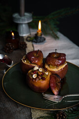 Three baked apples stuffed with nuts, raisins and lingonberries on big green plate  on table decorated with candles, cones, fir branches on dark background .