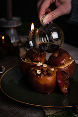 Pouring syrup on one of three baked apples stuffed with nuts, raisins and lingonberries on big green plate  on table decorated with candles, cones, fir branches on dark background .