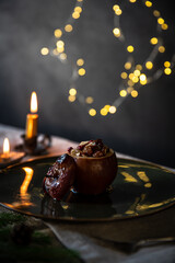 One baked apple stuffed with nuts, raisins and lingonberry, poured with syrup on big green plate with candle and saucer with fresh lingonberries and fir branches on wooden table.
