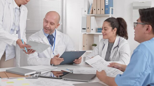 Over shoulder of unrecognizable person sitting at table with male Caucasian and female Mixed-Race colleagues working with papers, talking, cropped doctor leaning down with clipboard for signature