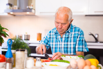elderly man cuts vegetables for salad at the table in the kitchen
