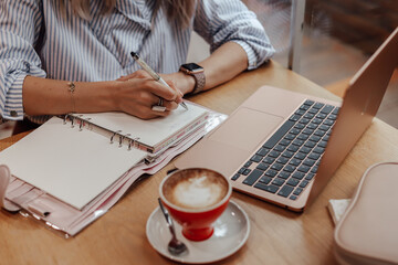 The cropped girl is working, making notes in the planner. Nearby is a cup of coffee and a laptop.