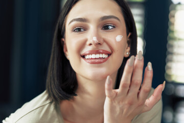 Happy girl with natural makeup applying cream on her face. Brunette young woman using moisturizing cream.