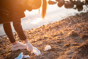 Young woman picking up disposable medical mask with sea and sunset at background.