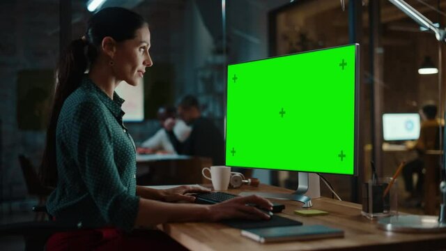 Middle Aged Multiethnic Specialist Working on Desktop Computer with Green Screen Mock Up Display in Busy Creative Office. Beautiful Diverse Female Manager in Green Polka Dot Blouse.