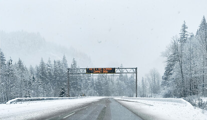 The road between tall spruce trees and mountain peaks in snowy weather. Road sign warning of dangerous road conditions. Coniferous snowy forest in the Rocky Mountains. British Columbia, Canada