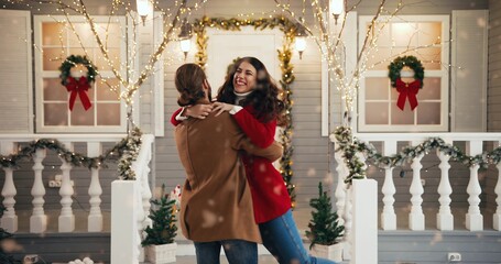 Happy Caucasian married couple in good mood outdoor in front of decorated home while snowing hugging and spinning on New Year Eve. Husband embracing his beautiful wife. Holidays concept Love relations