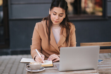 Attractive young business woman writing down something in notebook. Female drink coffee and working outdoors using laptop