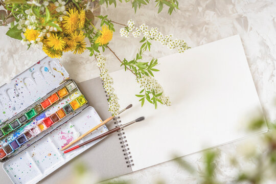 Top view of a table with a sketchbook, paints and brushes next to a bouquet of spring flowers