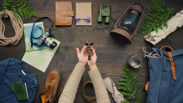 Top view flat lay shot of unrecognizable woman opening compass and spinning arrow. Travel accessories lying on wooden table decorated with birch logs and fern