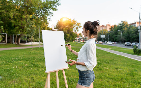 woman artist city summer, background car road, green lawn, begins paint picture on white canvas, hand brush color palette of paints, easel. Clothes white shirt shorts. Hair bun. Free space side view. - Powered by Adobe