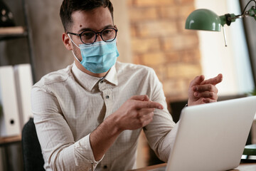 Young businessman with a mask working in his office on laptop. Businessman having video call. COVID - 19 virus protection.