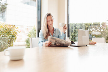 Enthusiastic beautiful young woman housewife reads her favorite newspaper in kitchen while drinking tea. Young woman is up to date with world news. Modern interior,large Windows overlooking the garden