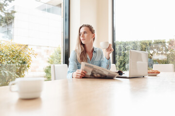 Enthusiastic beautiful young woman housewife reads her favorite newspaper in kitchen while drinking tea. Young woman is up to date with world news. Modern interior,large Windows overlooking the garden