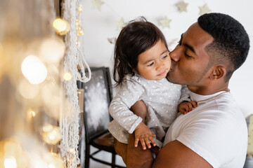 Positive young African American father holds in his arms charming mixed race girl in and looks at warm white lights. Positive loving father looks with his cute daughter at Christmas lights