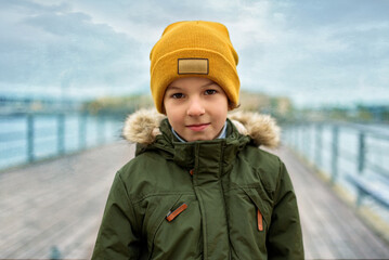 A boy dressed in an orange hat and green jacket stands against a gloomy sky.