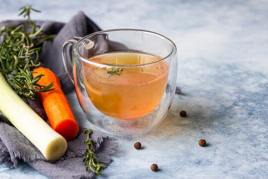 Homemade bone broth in glass mug and vegetables, blue concrete background. Collagen source for the body. Selective focus.