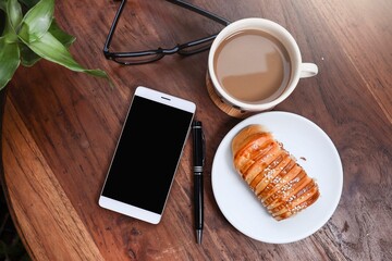 Top view of a smartphone on the wooden garden table.   A pen, red bean bread, a cup of coffee, and eyeglasses 