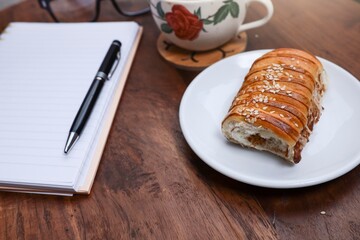 Selective focus on the red bean bread on the white plate with a pen, notebook, a cup of coffee, and eyeglasses on the wooden garden table 