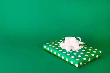 A beautifully wrapped gift with a white bow lies on a green background. The gift box is packed in green paper with white peas. Minimal Christmas New Year concept