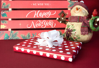 Festive background with the inscription "Happy New Year". A gift box with a large white bow lies on a red background. On the right is a snowman in a red knitted scarf and a hat