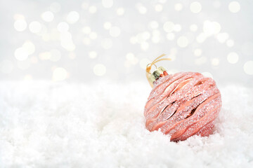 A shiny rose gold Christmas decoration lies on white snow. Glass decoration for a Christmas tree on a white background. Bokeh in the background. Close-up