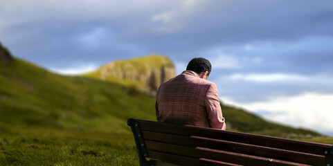 Fototapeta premium A lone man sits on a bench beside a green summer mountain.