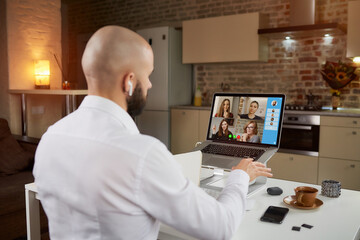 Back view of a male employee in earphones who is explaining and gesticulating on a business video conference on a laptop. A bald man with a beard on an online meeting with his colleagues at home