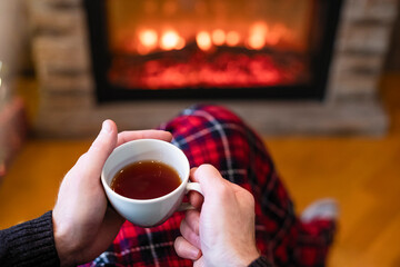 Man with cup coffee, tea in santa claus hat sitting and warming at winter evening near fireplace...