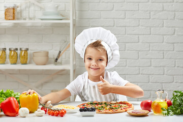 little boy in chef hat and an apron cooking pizza in the kitchen. child showing Ok gesture. delicious