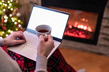 Christmas. Man with laptop sitting near christmas tree and fireplace with cup of tea. Concept of online service in holidays. Back view.