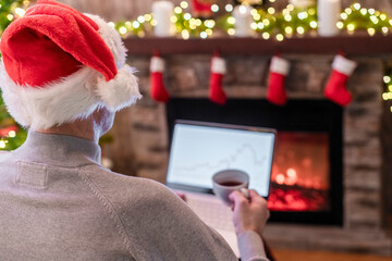 Man freelancer in santa claus hat working on laptop with graphs and charts on screen sitting near christmas tree and fireplace.