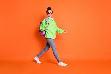 Fototapeta na wymiar Full length photo of adorable walking woman wear green hoodie schoolbag glasses isolated vivid orange color background