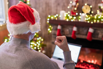 Back view of angry man freelancer in santa claus hat, raising and swinging fist on laptop screen sitting near christmas tree and fireplace. Concept