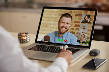 A laptop screen view of the telecommunications application during an online meeting over a man's shoulder. Back view of an employee who is holding earphones on a video conference with colleagues.