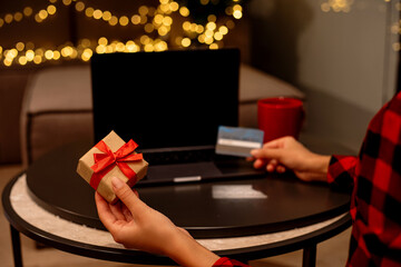 A woman holds a small red gift box in her hands and uses a credit card to search on a laptop.  