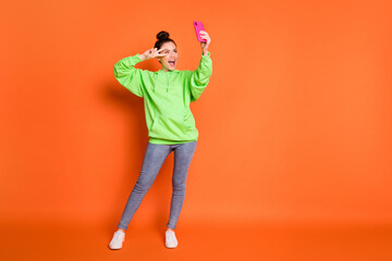 Fototapeta na wymiar Full length photo portrait of girl making v-sign near eye holding phone in one hand laughing taking selfie isolated on vivid orange colored background